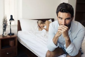 Tristezza post orgasmo: nell'immagine un uomo seduto sul letto con aria malinconica. La sua partner lo osserva in secondo piano, sotto le lenzuola