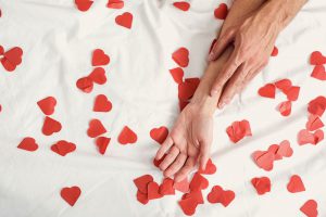 Cose da non fare durante il rapporto sessuale: nell'immagine si vede una mano maschile appoggiata su di una mano femminile su di un letto cosparso di cuori rossi di carta.